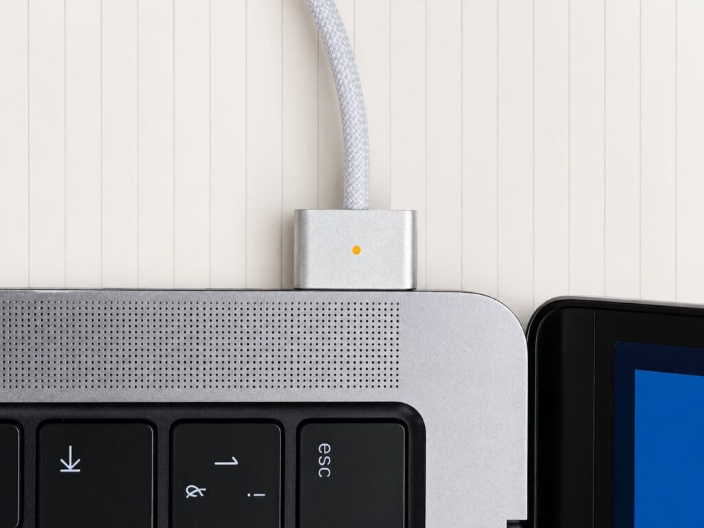 Gros plan du connecteur MagSafe Apple branché sur un MacBook avec voyant lumineux orange indiquant une charge en cours