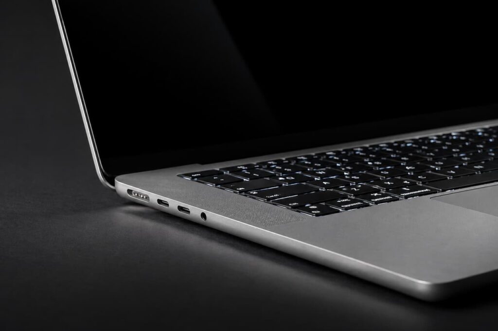 Gros plan du côté gauche d'un MacBook Pro argenté avec port MagSafe, ports USB-C Thunderbolt, clavier rétroéclairé allumé et écran noir sur fond sombre