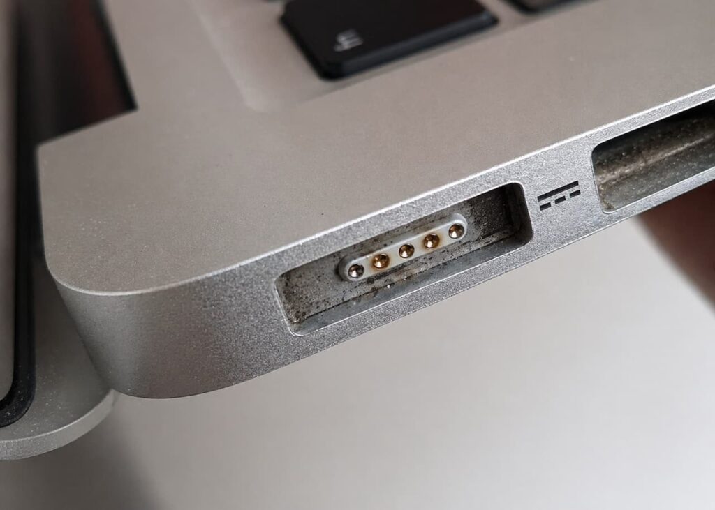 Gros plan du port MagSafe d'un MacBook encrassé avec accumulation de poussière et débris sur les broches dorées et autour de l'entrée de charge
