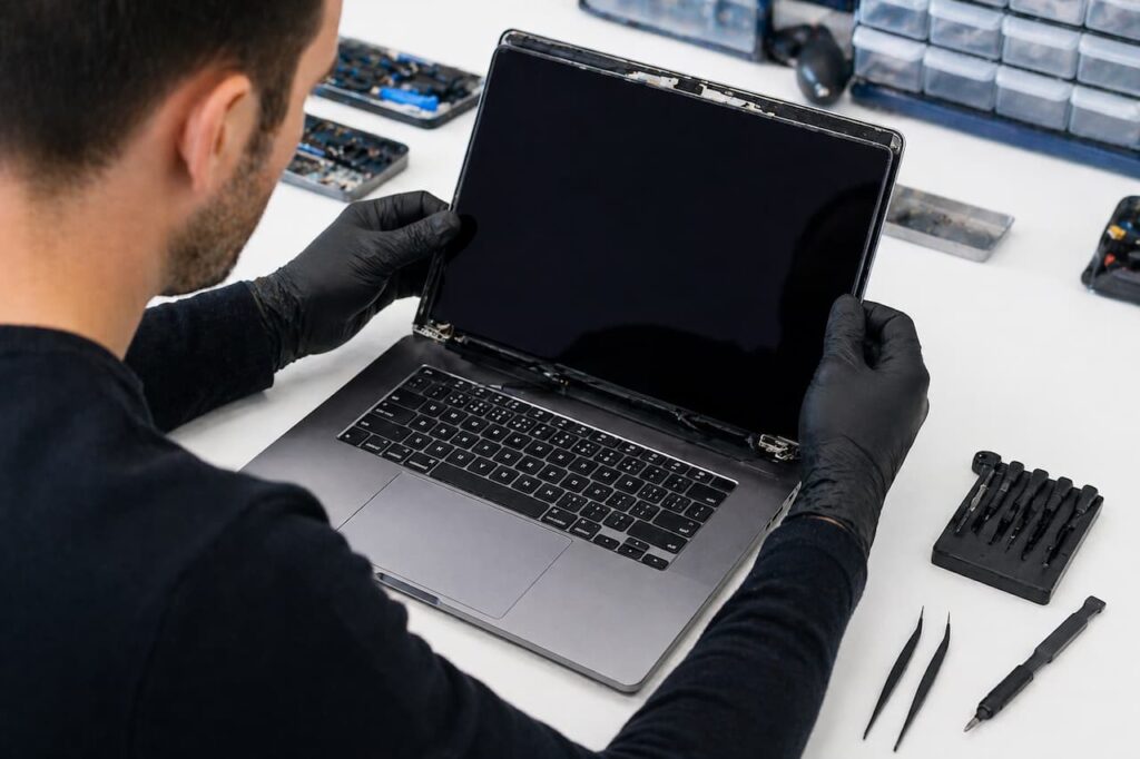 Technicien avec gants noirs en train de démonter et remplacer la dalle d'un MacBook Pro gris sidéral sur un établi blanc avec outils de précision