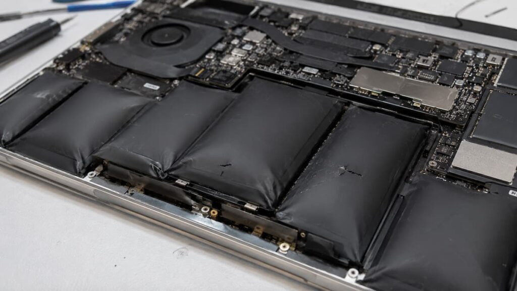 Gros plan de l'intérieur d'un MacBook Pro ouvert posé sur une surface blanche, révélant une batterie multi-cellules fortement gonflée avec des cellules noires visiblement bombées et déformées, carte mère et ventilateur visibles en arrière-plan, outils de réparation posés à gauche