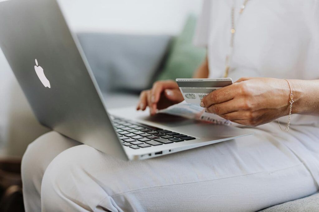 Femme en tenue blanche tenant une carte bancaire dans une main et tapant sur le clavier d'un MacBook Air argenté posé sur ses genoux, dans un intérieur lumineux